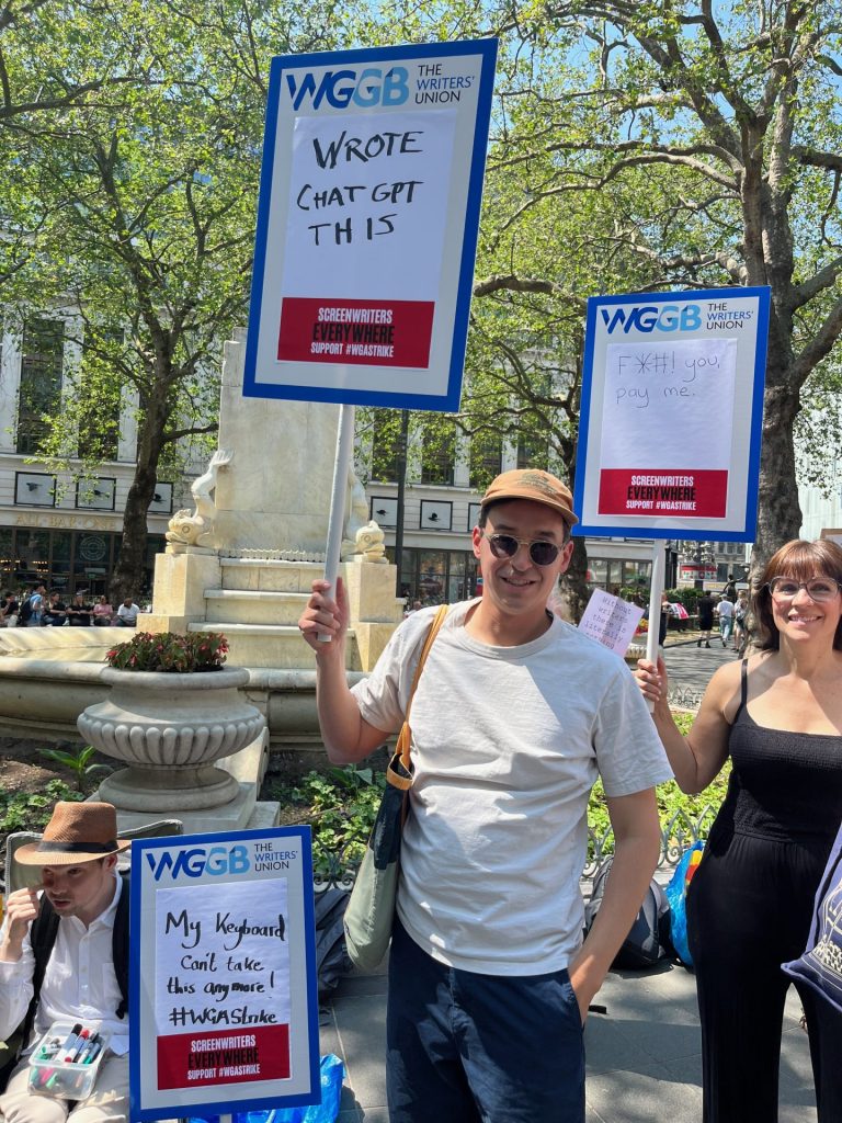 Writer Andrew Chambliss at a protest in support of Writers' Guild of America strikes. He is holding a Writers Guild of Great Britain Sign reading 'Wrote chat GPT this'. He is wearing a white t-shirt, navy-blue trousers, sunglasses and a brown baseball cap. Other protesters and signs are visible in the background. 