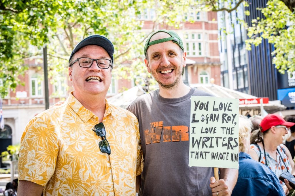 'Doctor Who' Writer Russell T Davies, and 'His Dark Materials' writer Jack Thorne  at the Writers' Guild of Great Britain protest in solidarity with Writers Guild of America Strikes. Davies is wearing glasses, a yellow floral shirt and a blue baseball cap. Thorne is wearing a grey t-shirt and green baseball cap while holding a sign which reads 'You love Logan Roy, I love the writers that wrote him.'