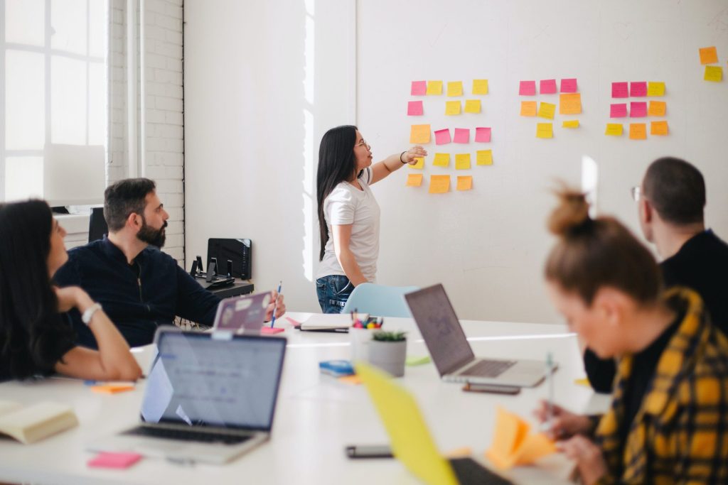 A woman pointing at post it notes on a white board, with people sat around a table listening to her