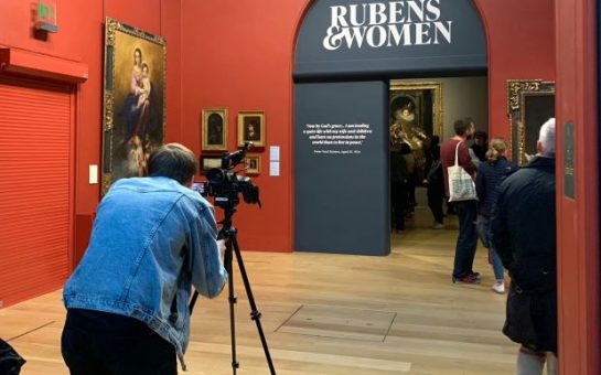 A man taking a picture of the entrance to the Rubens and Women exhibition at Dulwich Picture Gallery. The walls are red, the entering arch blue and people are littered about. Pictures adorn the walls.