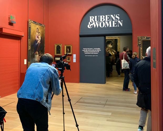 A man taking a picture of the entrance to the Rubens and Women exhibition at Dulwich Picture Gallery. The walls are red, the entering arch blue and people are littered about. Pictures adorn the walls.