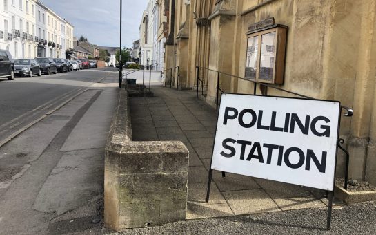 A white sign reads "polling station" outside an old community building next to a road.