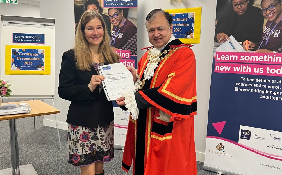 Oana-Maria-Vasile, adult learner accepting certificate from Mayor of Hillingdon in front of Adult Education posters in council offices.