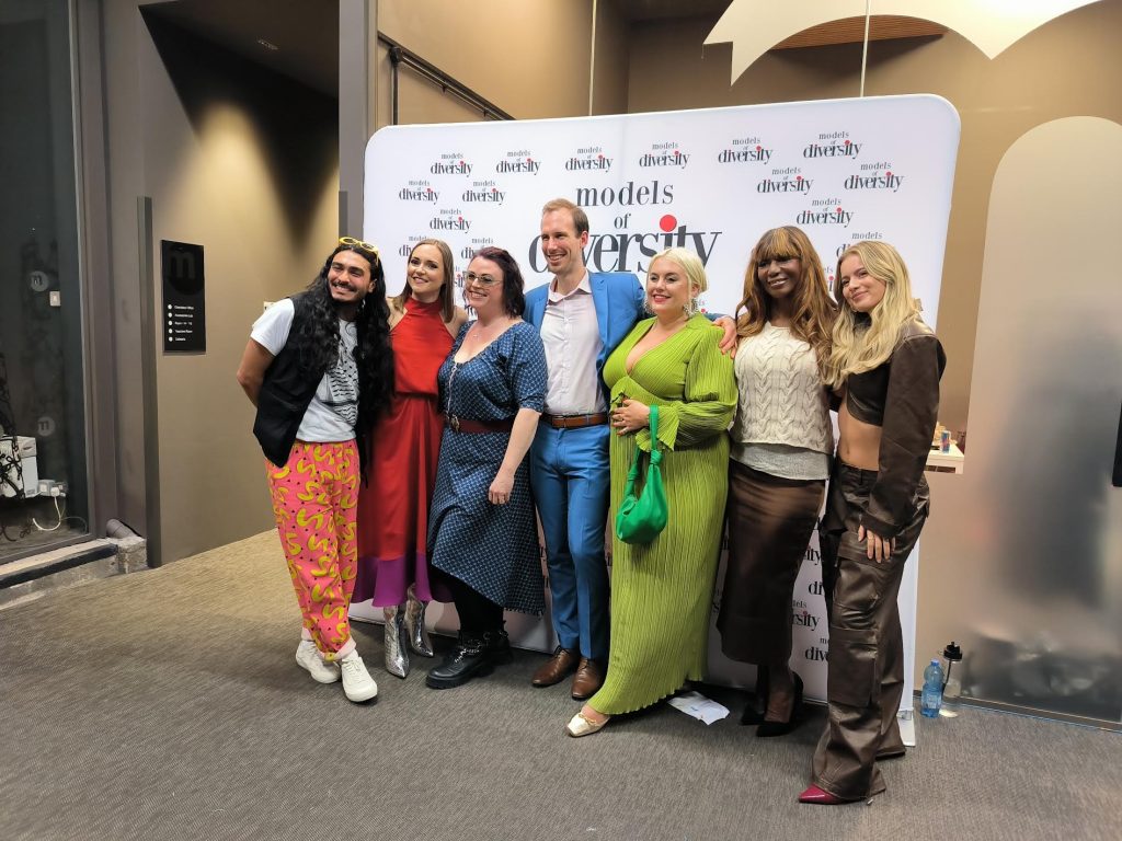 Image shows seven panel speakers posing for a photo in front of a white backdrop with black writing that reads 'Models of Diversity'. 