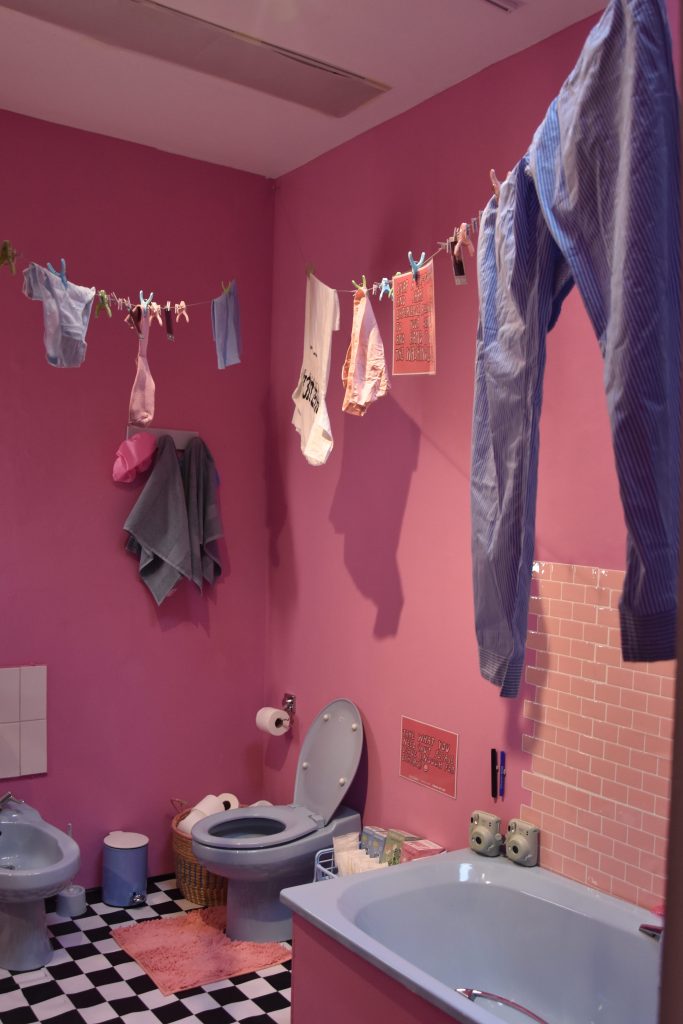 A view of a bathroom with pink walls and light blue fixtures, with two instant cameras on the corner of the tub and an assortment on sanitary products on the side of the toilet.