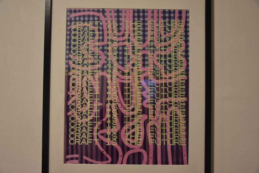 A tapestry artwork made of three different patterns, in the colours of violet, purple and pink, with the phrase "Craft is the future" repeatedly embroidered across it in yellow. The artwork is enclosed in a black frame hanging on a white wall.