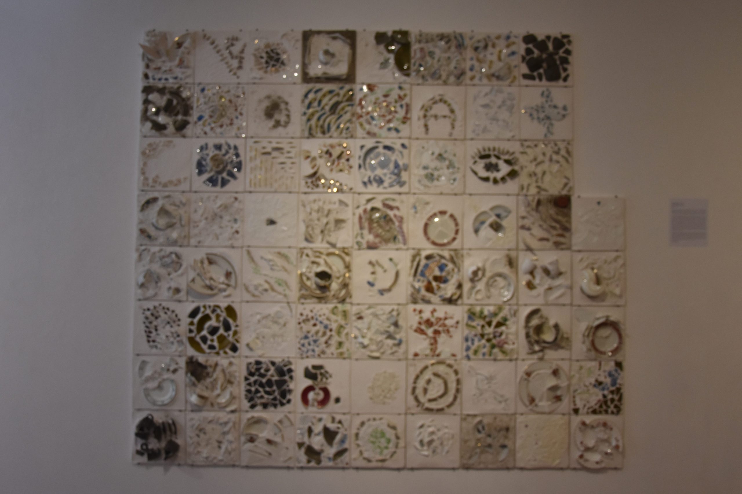 A white plaster panel hanging on a white wall, comprised of 69 tiles made of broken crockery.