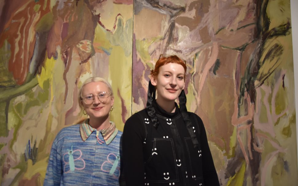 Co-founders Eliza Hatch (left) and Bee Illustrates (right) pose smiling in front of an artwork at the 2024 Hysterical: Radical Creativity exhibition in Bermondasey, London