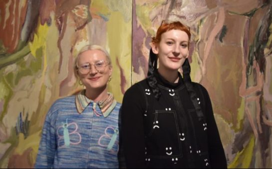 Co-founders Eliza Hatch (left) and Bee Illustrates (right) pose smiling in front of an artwork at the 2024 Hysterical: Radical Creativity exhibition in Bermondasey, London