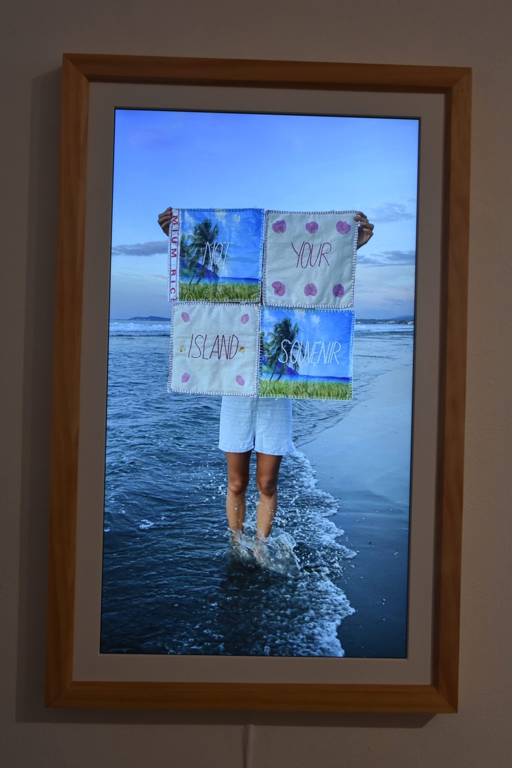 A framed picture hanging from a white wall of a woman standing on a beach with her feet in the water and holding up a tapestry with the words "Not your island souvenir" embroidered on it.