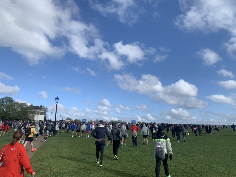 Runners and supporters walking towards the London Marathon starting line across a field on a sunny day.