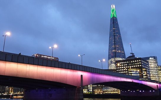 Picture of London Bridge, lit at night, from the lower, northern side of the river. The Shard is in the background to the upper-right.