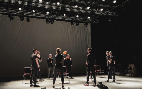 People in a circle on a stage wearing all black and passing a ball around the circle.