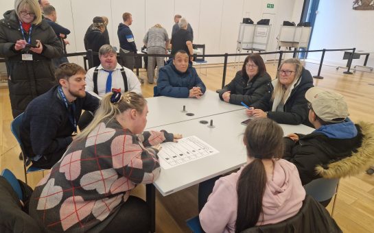 Group of people with learning disabilites sit around a table and learn about the voting process