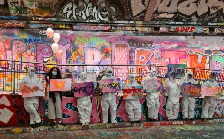 Nine children lined up holding spray painted signs in front of a graffiti covered wall.