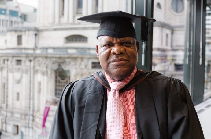 Chris Osagbaekhoe stands by a window in academic dress for his graduation