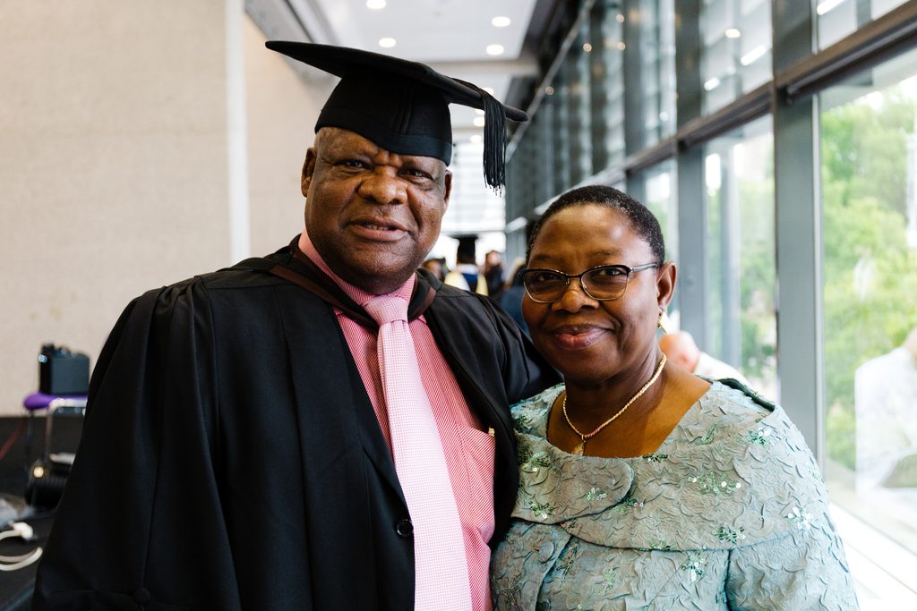 Chris Osagbaekhoe standing with his wife, Ayo Osagbaekhoe. Chris is dress in a cap and gown and his wife is wearing a blue dress.