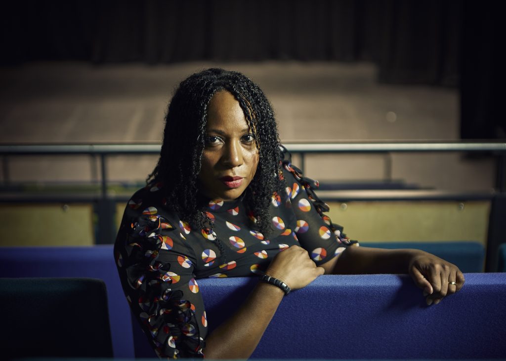 Dr Suzann McLean is sitting on blue theatre chairs facing backwards with her right arm over the back of the chair. She is looking towards the camera with a serious expression and wearing a black top with colourful circles.